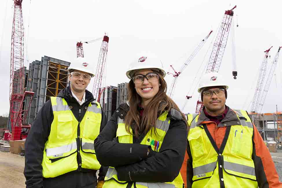 Two men and a woman standing with hardhats, vests, and goggles in front of cranes and other equipment at Stonewall construction site.
