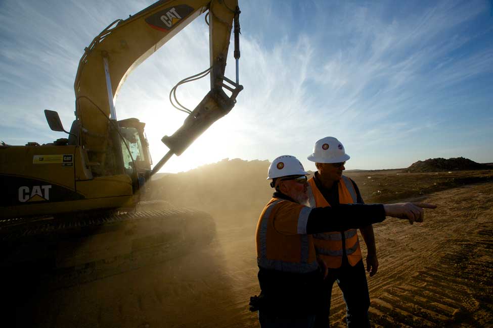 two workers consult each other with an earth mover in the background