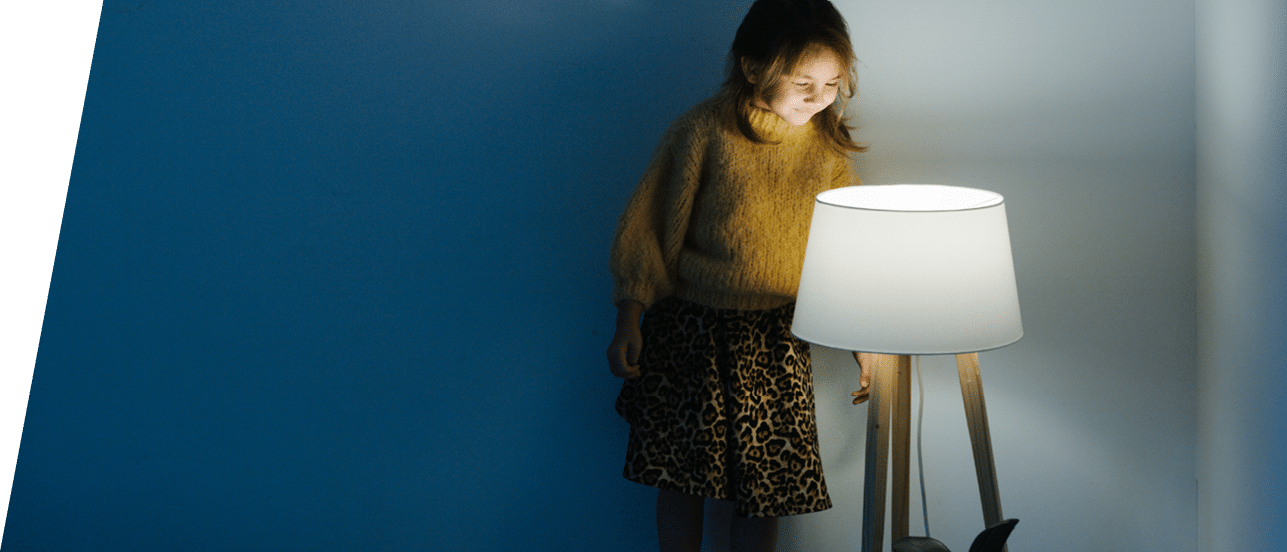 Brunette little girl in yellow knitted sweater and leopard skirt standing on a backseat of a couch at home  She's looking at dim night light from above  Her face is lit 